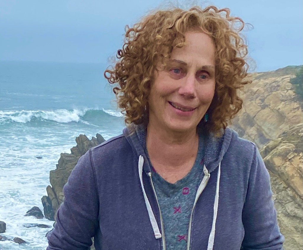 woman with blonde curly hair and blue sweatshirt in front of cliffs and ocean
